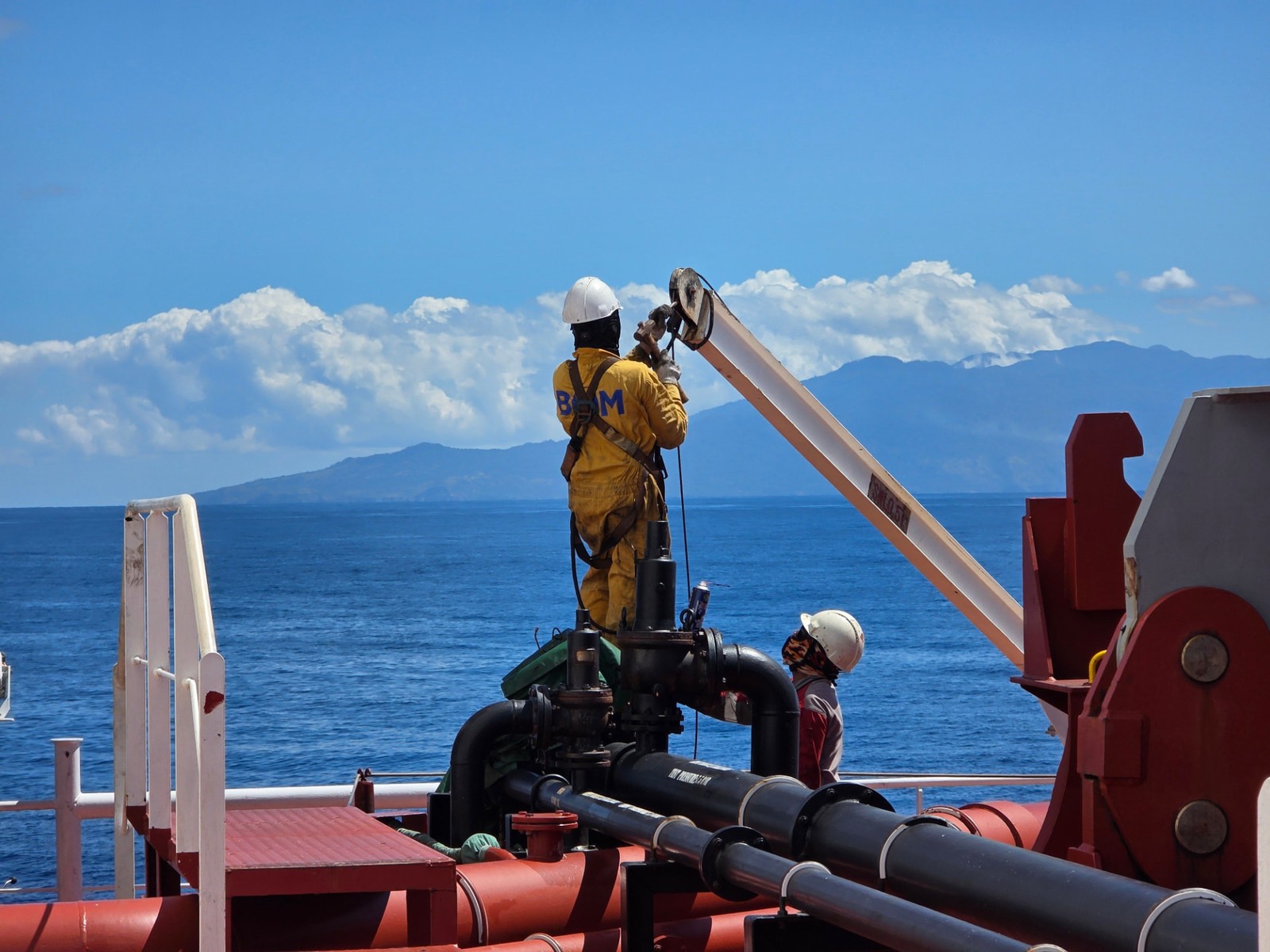 Offshore personnel operating deck equipment on a vessel with open sea and coastline in the background